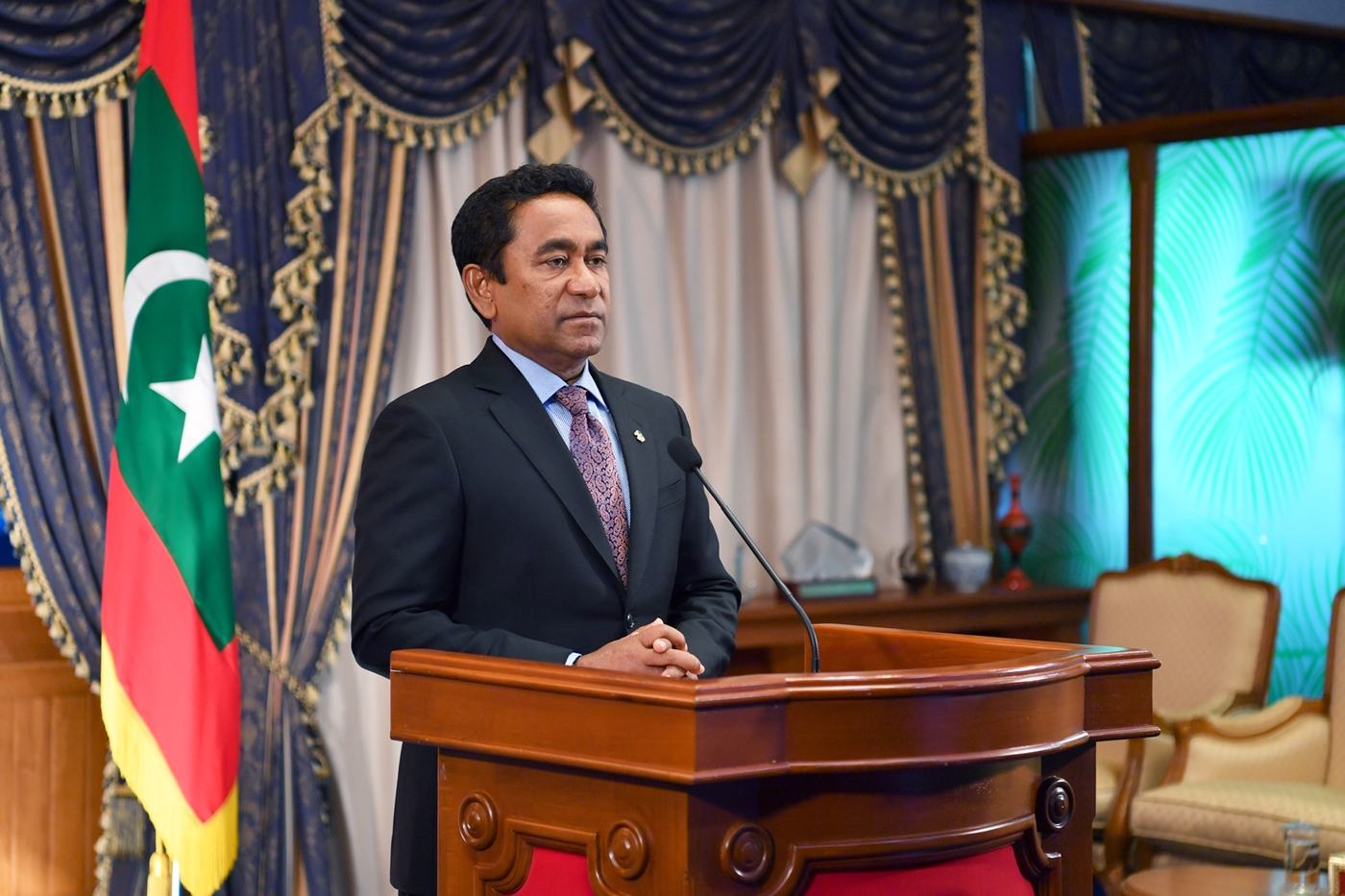 Abdulla Yameen speaking at a podium beside the national flag of the Maldives.